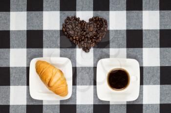 Cup of coffee, croissant and heart made of coffee beans on black and white checkered tablecloth. Top view.