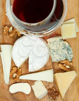 top view of red wine glass and assortment of cheeses on wooden plate close up