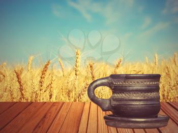 Old black tea mug on wooden surface against of golden wheat ears and blue sky.