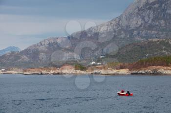 Coastal Norwegian landscape with Mountains, sea and cloudy sky. Small red fishing boat is floating on water