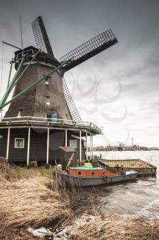 Windmill under dark cloudy sky on Zaan river coast, Zaanse Schans town, popular tourist attractions of the Netherlands. Suburb of Amsterdam