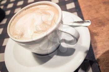 White cup of cappuccino stands on wooden table, retro style tonal correction filter and selective focus