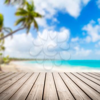 Empty wooden pier background over blurred tropical beach coastal landscape with palm tree, cloudy sky and bright sea water