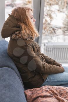Blond Caucasian teenage girl in warm clothes sitting on sofa near the window in hallway