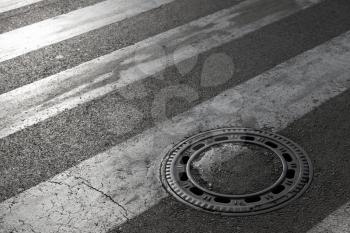 Sewer manhole cover on asphalt road with pedestrian crossing marking