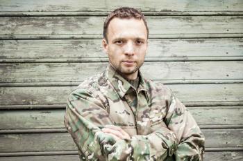 Young serious Caucasian military man in camouflage uniform with crossing hands. Closeup portrait over green wooden wall
