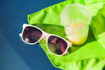 Sunglasses and plastic glass of juice stand on green towel over blue sun lounger, summer beach resort vacation theme