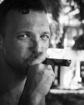 Young European man smokes big cigar, close up black and white portrait with selective focus. Dominican Republic