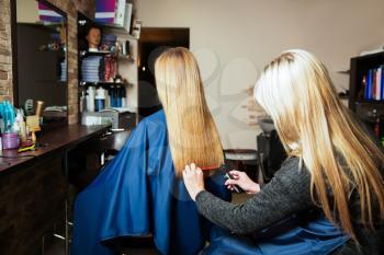 Professional hairdresser with machine for hair and hairbrush in hands behind young female in hairdressing salon.