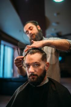 Bearded hairdresser making male hipster hairstyle. Client man in black salon cape visit barbershop