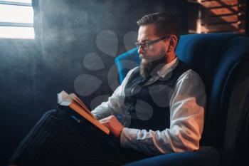 Portrait of bearded man in glasses sitting in armchair against window with sunlight in smoky room. Writer, journalist, literature author, blogger or poet concept