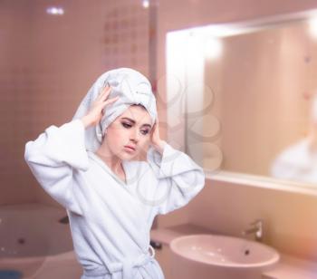 Beautiful woman in bathrobe with towel on head take care of her hair against mirror in bathroom