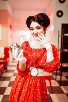 Surprised pin up girl looks at the alarm clock, vintage cafe interior on background, popular american fashion 50s and 60s. Red dress with polka dots, bright make-up