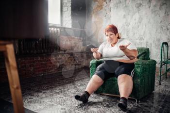 Fat woman sits in a chair and watches TV. Unhealthy lifestyle, obesity