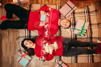 Happy love couple with gifts lies on the floor, top view, christmas holidays. Xmas celebration, cheerful man and woman in red pullovers, fir-tree with decoration on background