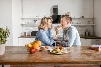 Love couple sitting at the table, romantic dinner. Man and woman drinks wine and eats fruits on the kitchen. Happy lifestyle, beautiful relationship