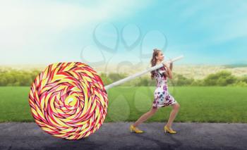 Young pretty girl drags a huge colorful candy. Bright girl with blonde curly hair. Stylish girl in colorful summer dress and yellow shoes, summer meadow on background.