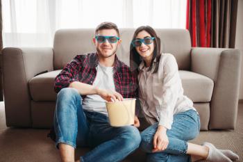 Smiling couple sitting on the floor against couch and watch tv with popcorn at home, man with remote control in hand, window on background