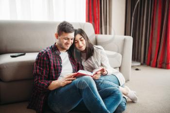 Happy love couple sitting on the floor and looks at the book together. Modern apartment interior of living room on background