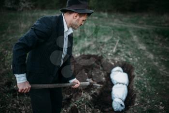 Maniac with a shovel buries victim into a grave, the body wrapped in a canvas, serial murderer concept