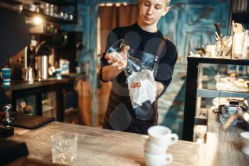 Young male barista wipes the dishes after making coffee at cafe counter. Barman works in cafeteria, bartender occupation