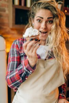 Housewife in apron eating sweet dessert with cream, kitchen interior on background. Female cook tastes fresh homemade cake. Chef eats pie