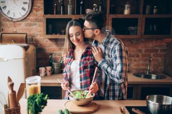 Young husband kissing his wife on the kitchen while she cooking breakfast. Man and woman preparing vegetable salad, happy family together