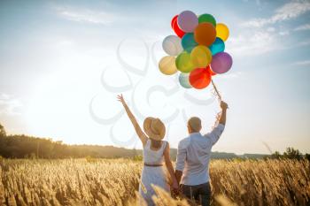Love couple with balloons, leisure in a rye field on sunset. Pretty wife and husband walking on summer meadow