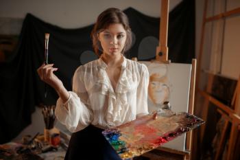 Female artist with color palette and brush standing against easel in studio. Creative paint, painter drawing portrait, workshop interior