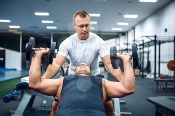 Athlete lies on bench and doing exercise with dumbbell under instructor control, motivation method, gym interior on background. Weightlifting workout in sport or fitness club