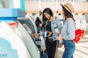 Two female travelers withdraw cash at ATM in airport. Passengers with baggage in air terminal, back view, happy journey of white and black ladies, summer travel