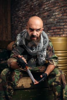 Terrorist in uniform with knife in hands sitting on boxes of ammunition. Terrorism and terror, soldier in camouflage in weapon arsenal, barrels of fuel or chemicals on background