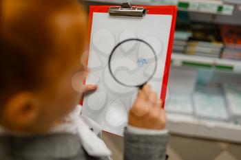 Little schoolgirl looks at the picture through magnifying glass, shopping in stationery store. Female child buying office supplies in shop, schoolchild in supermarket