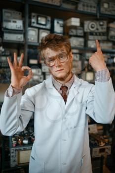 Strange male scientist shows signs with his fingers in laboratory. Electrical testing tools on background. Lab equipment, engineering workshop