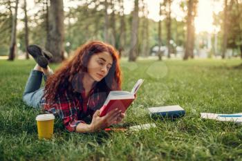 Female student with cup of coffee lying on the grass in summer park. Ginger teenager studying and leisures outdoors