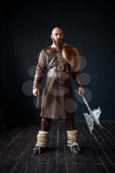 Viking with axe dressed in traditional nordic clothes. Ancient warrior, studio with black walls and wooden floor on background
