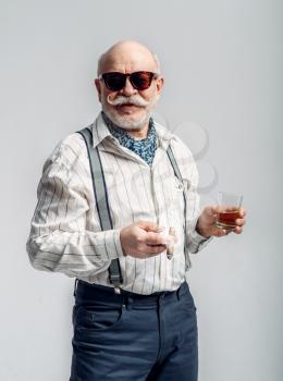 Fashionable elderly man with cigar and bottle of good alcohol, grey background. Mature senior looking at camera in studio, dude