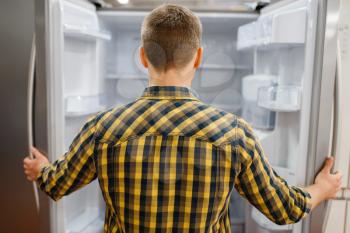 Young man at the opened refrigerator in electronics store. Male person buying home electrical appliances in market