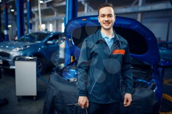 Repairman in uniform stands at vehicle with opened hood, car service station. Automobile checking and inspection, professional diagnostics and repair