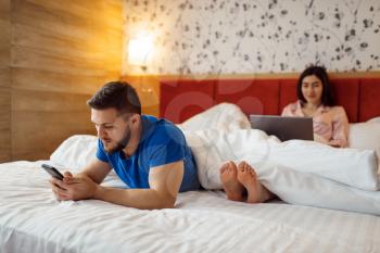 Love couple uses their gadgets in bed, good morning. Harmonious relationship in young family. Man and woman resting together in their house, carefree weekend