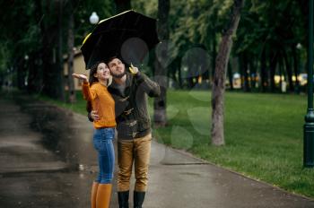 Happy love couple enjoys summer rainy day. Man and woman stand under umbrella in rain, romantic date on walking path, wet weather in alley
