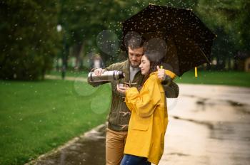 Love couple drinks hot tea in park, summer rainy day. Man and woman stand under umbrella in rain, romantic date on walking path, wet weather in alley