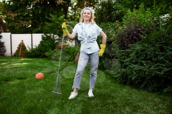 Smiling woman with rake works in the garden. Female gardener takes care of plants outdoor, gardening hobby, florist lifestyle and leisure