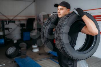 Auto repairman in uniform holds two new tires, repairing service. Worker repairs car tyre in garage, professional automobile inspection in workshop