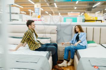 Young love couple sitting on beds in furniture store. Man and woman looking samples for bedroom in shop, husband and wife buys goods for modern home interior