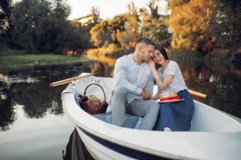 Love couple with fruit basket, relaxing in boat on quiet lake at summer day. Romantic date, boating trip, man and woman walking along the river