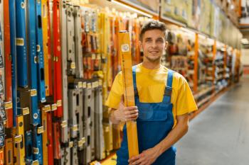 Male builder with building level in hardware store. Constructor in uniform look at the goods in diy shop