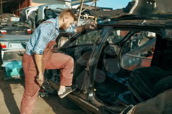 Male mechanic disassembles the car on junkyard. Auto scrap, vehicle junk, automobile garbage. Abandoned, damaged and crushed transport, scrapyard