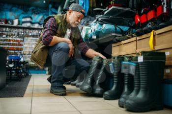 Male angler choosing rubber boots in fishing shop. Equipment and tools for fish catching and hunting