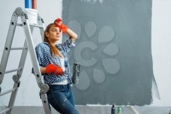 Tired female house painter with roller paints wall in grey color. Home repair, laughing woman doing appartment renovation, room decoration renovating